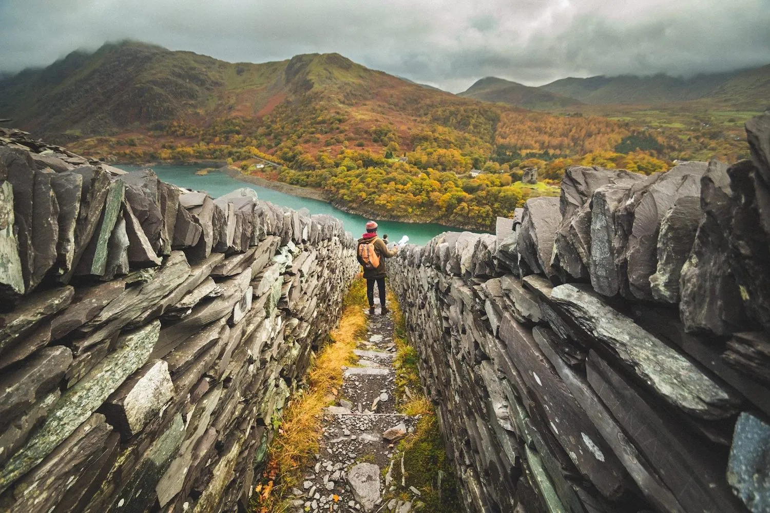 man standing between two high stone walls, surrounded by grassy hills & mountains